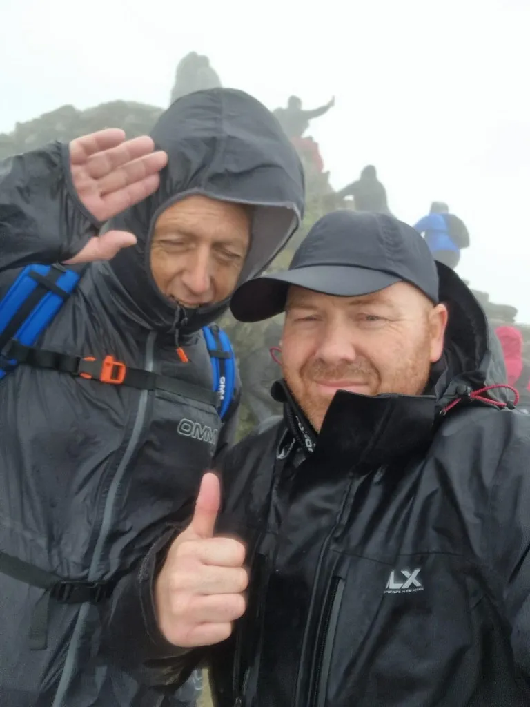 Two Mantech team members giving a thumbs up in heavy rain near a summit