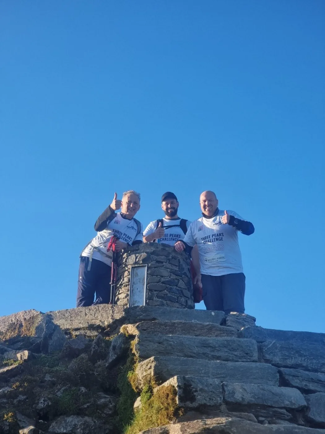 Three climbers in Mantech Three Peaks Challenge shirts giving thumbs up beside a stone trig point under a clear blue sky