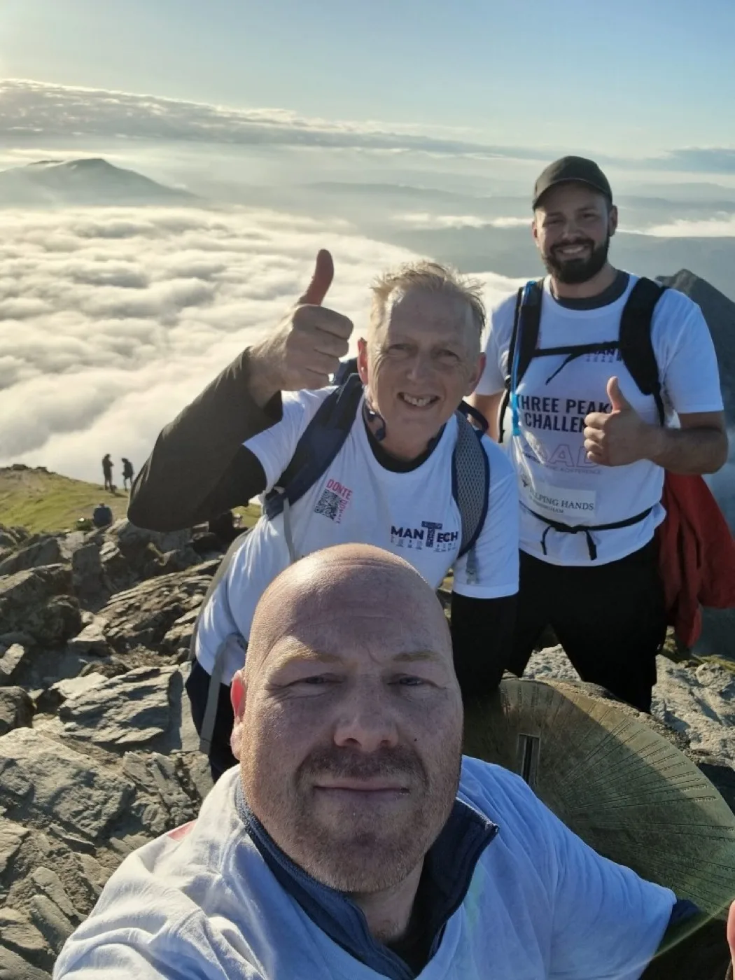 Mantech team at a Three Peaks summit above a cloud inversion — thumbs up in branded challenge shirts