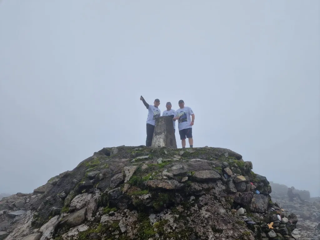 Three challenge participants standing at a misty mountain trig point