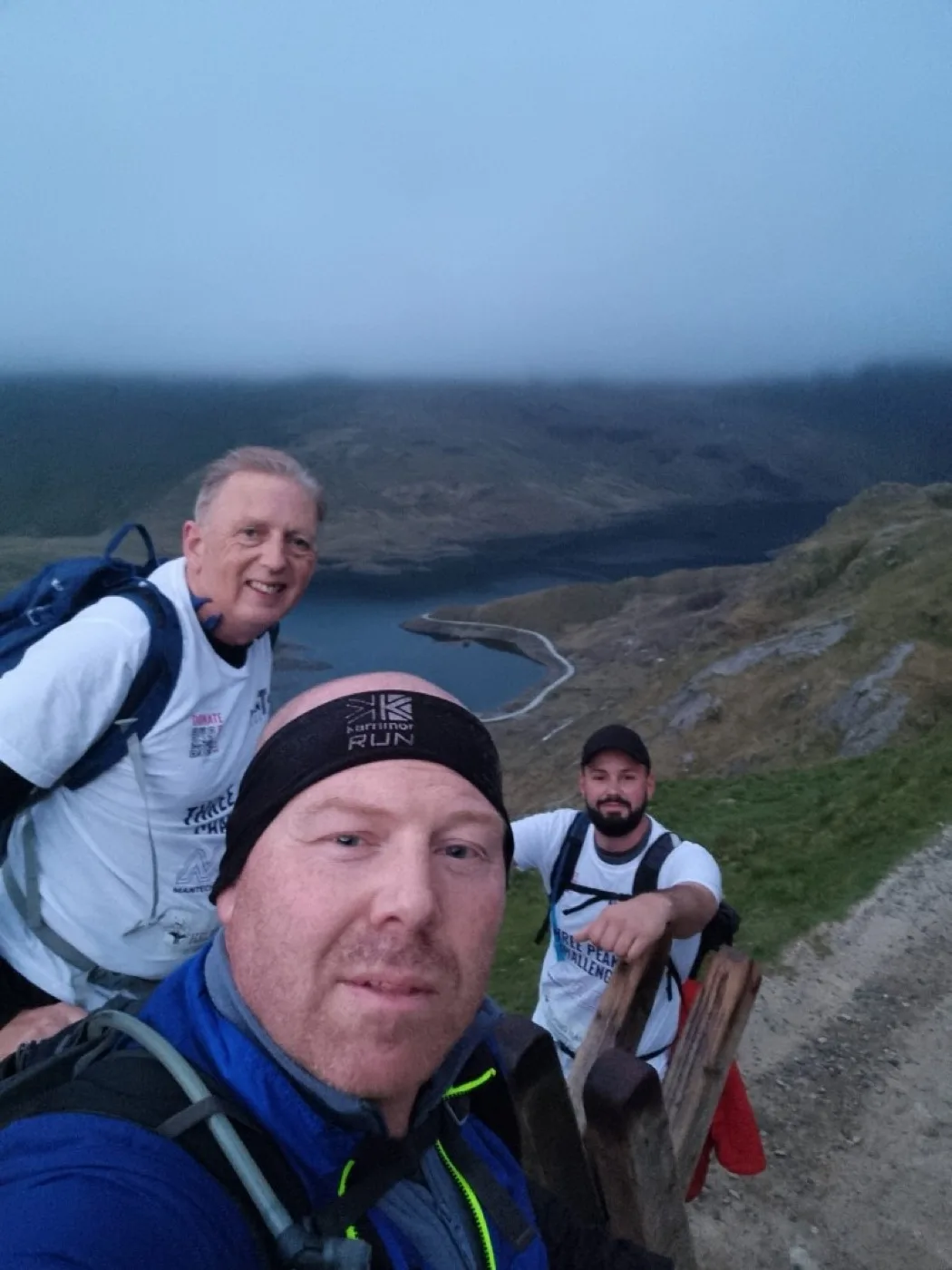 Three hikers on a mountain trail above a misty valley lake during the Three Peaks Challenge